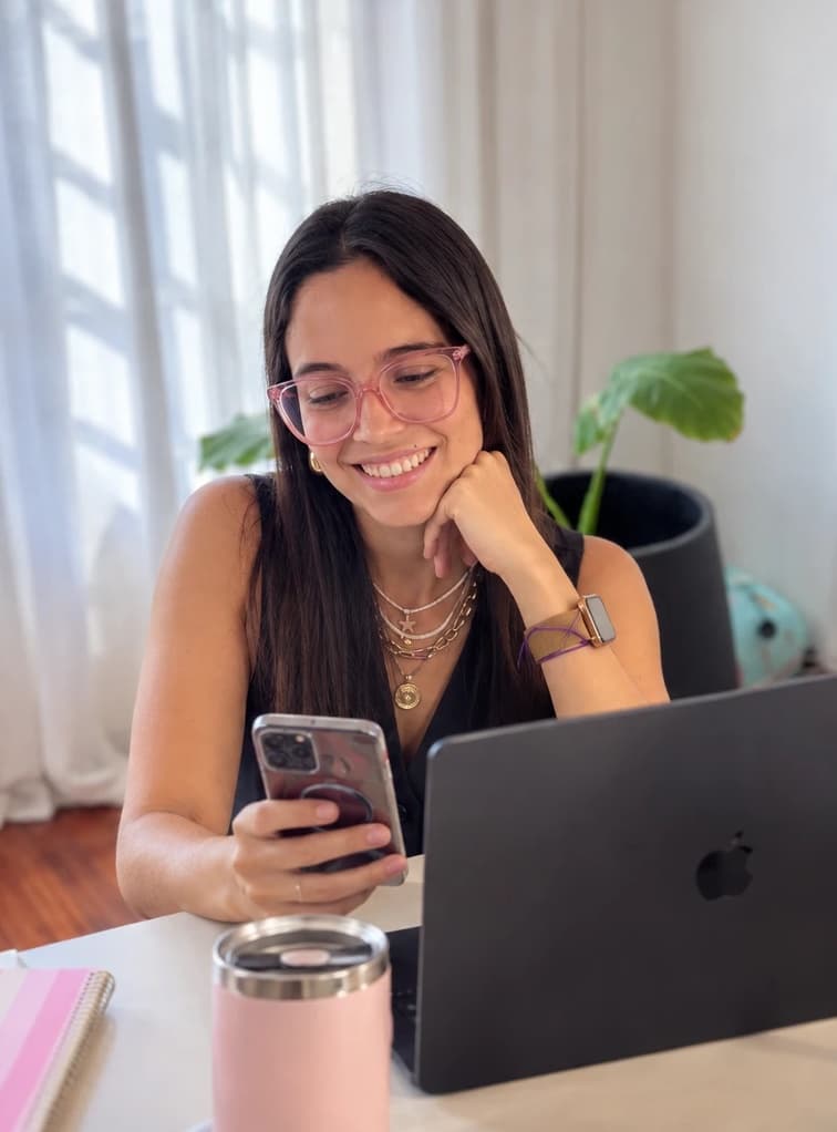 Julieta sonriendo mientras mira su celular, sentada frente a su computadora